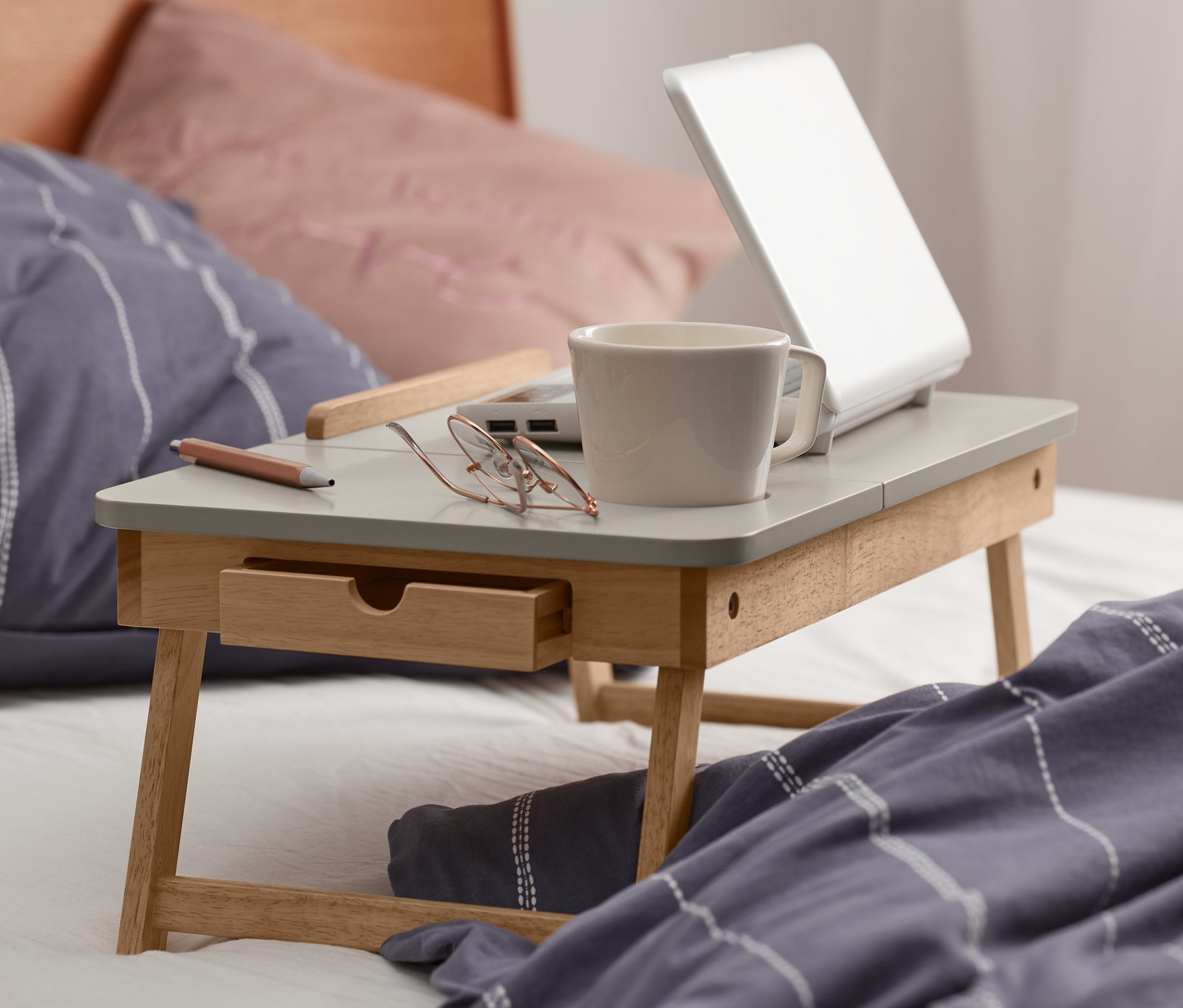 Betttisch mit Laptop, Tasse, Brille und Stift steht auf einem Bett.