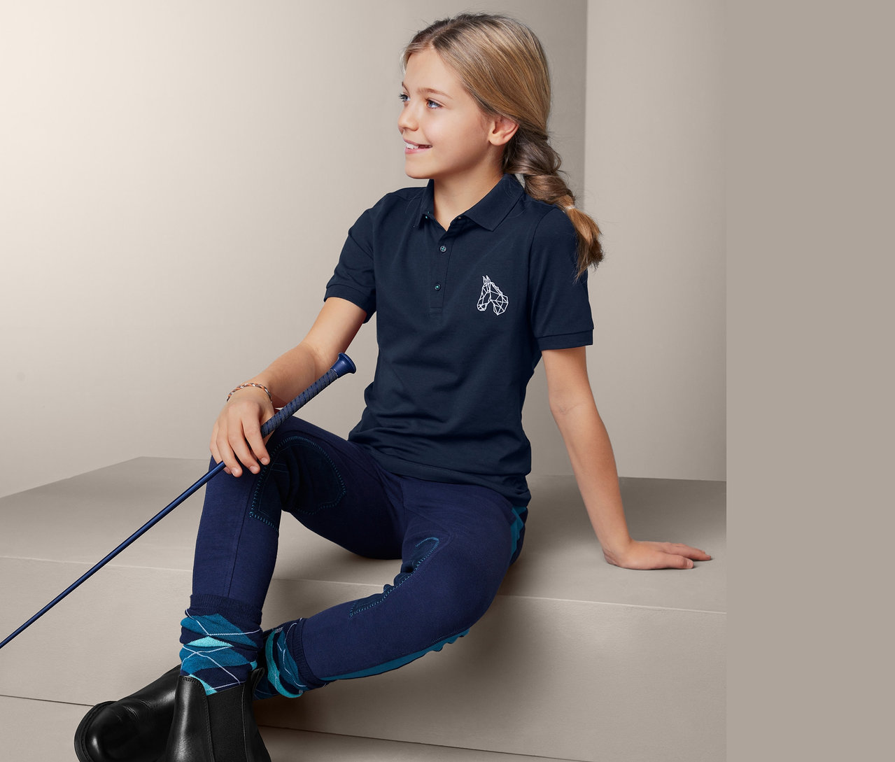 Mädchen sitzt und hält eine Reitpeitsche. Sie trägt ein Polohemd aus dem 2 Poloshirts Set.