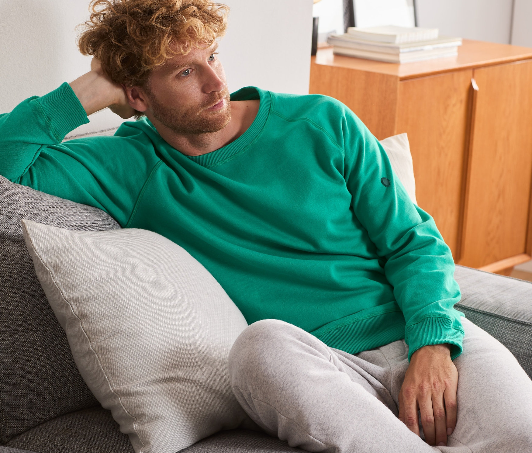 Mann liegt auf einem grauen Sofa und trägt ein grünes Sweatshirt.