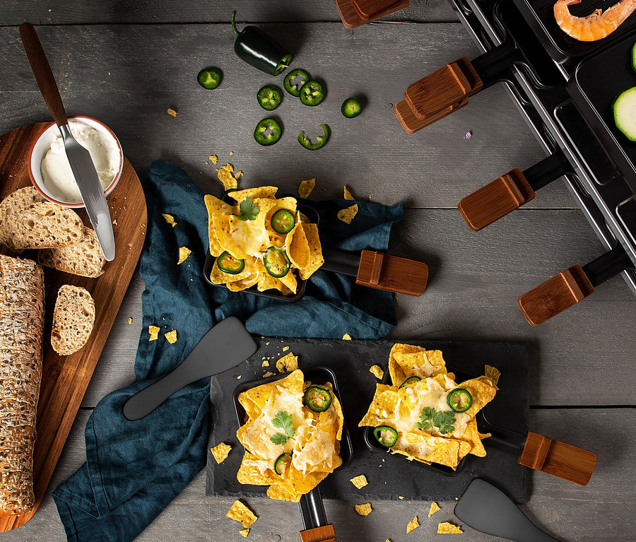 Raclettepfännchen mit Nachos, Käse und Jalapeños auf einem Tisch, daneben Brot, Käse und Gemüse. Im Hintergrund ist ein Raclettegrill zu sehen.