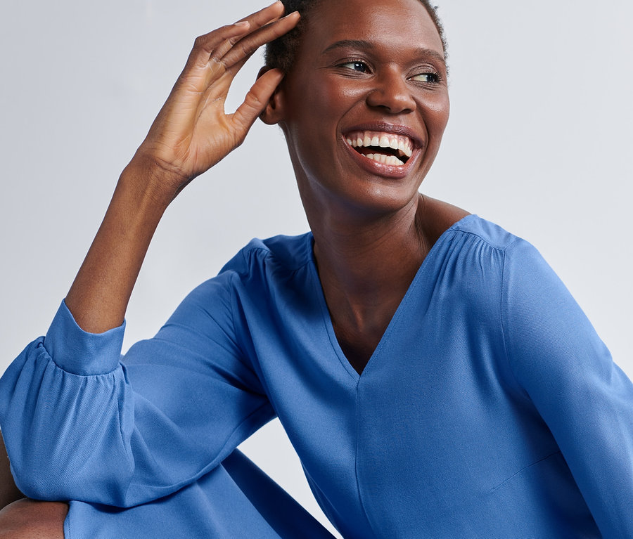 Lächelnde Frau berührt ihr Gesicht und posiert in einem Midikleid mit Gürtel.