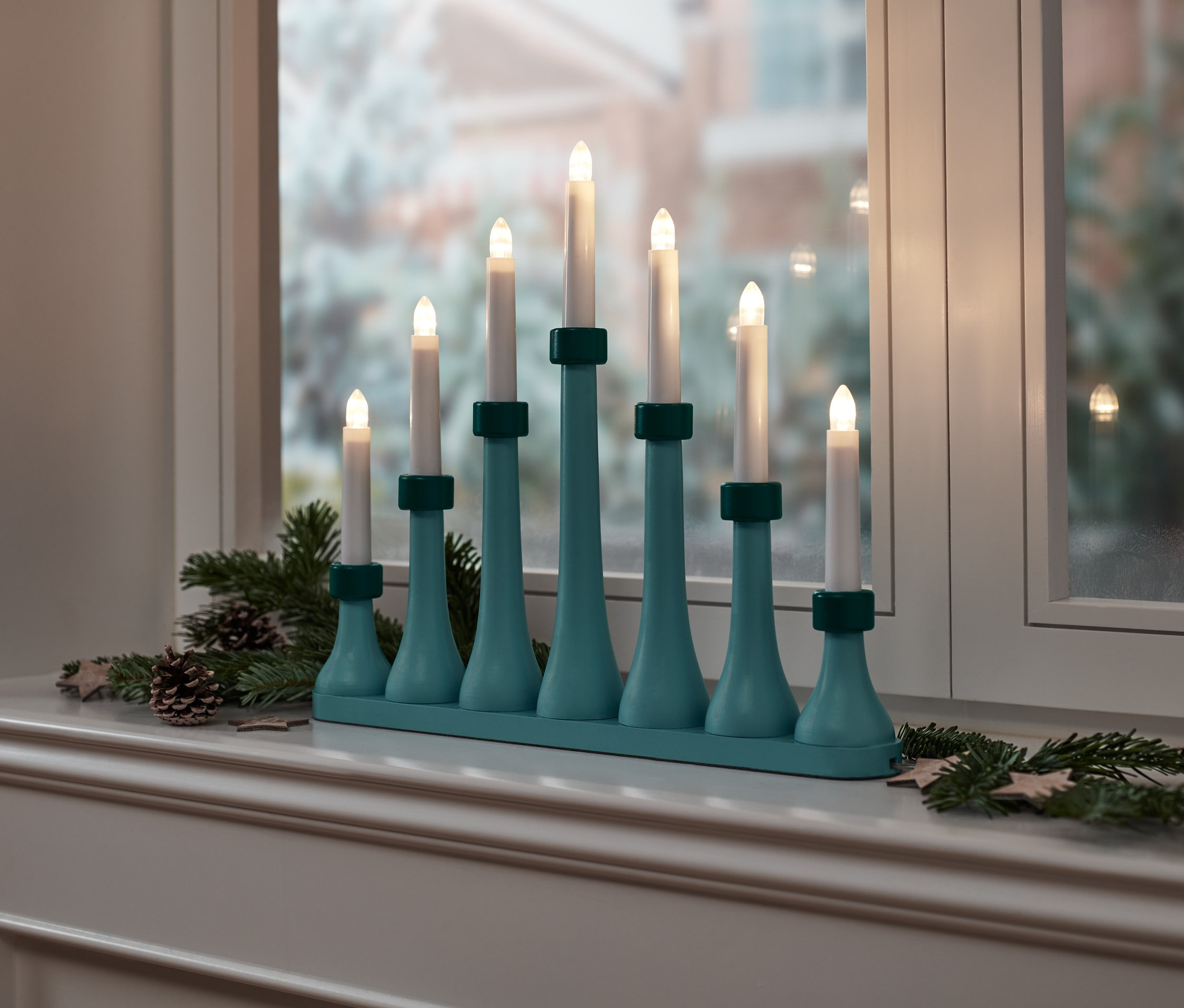Türkisgrüne LED-Kerzenpyramide steht auf einem Fensterbrett, dekoriert mit Tannenzweigen und Tannenzapfen.