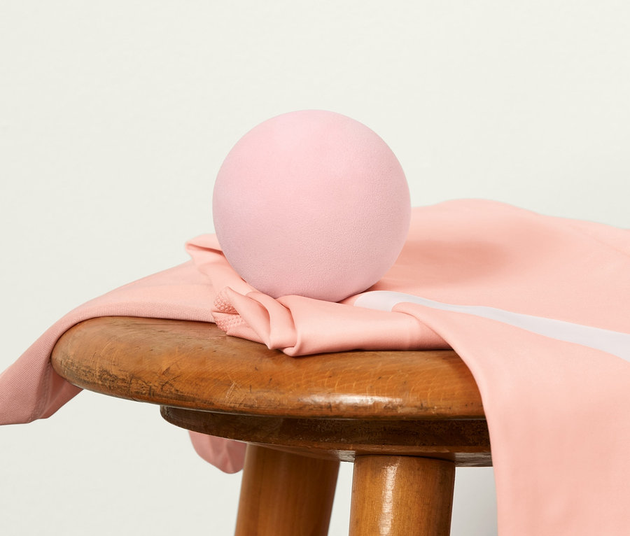 Ein pinker Faszienball liegt auf einer pinken Leggings mit weißem Streifen, die auf einem Holzhocker liegt. Im Hintergrund befindet sich eine pinke Yogamatte.