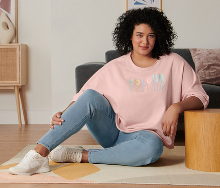 Eine Frau sitzt im Wohnzimmer auf dem Boden und trägt ein roséfarbenes Sweatshirt mit 3/4-Arm, Treggings und Veloursleder-Sneaker.
