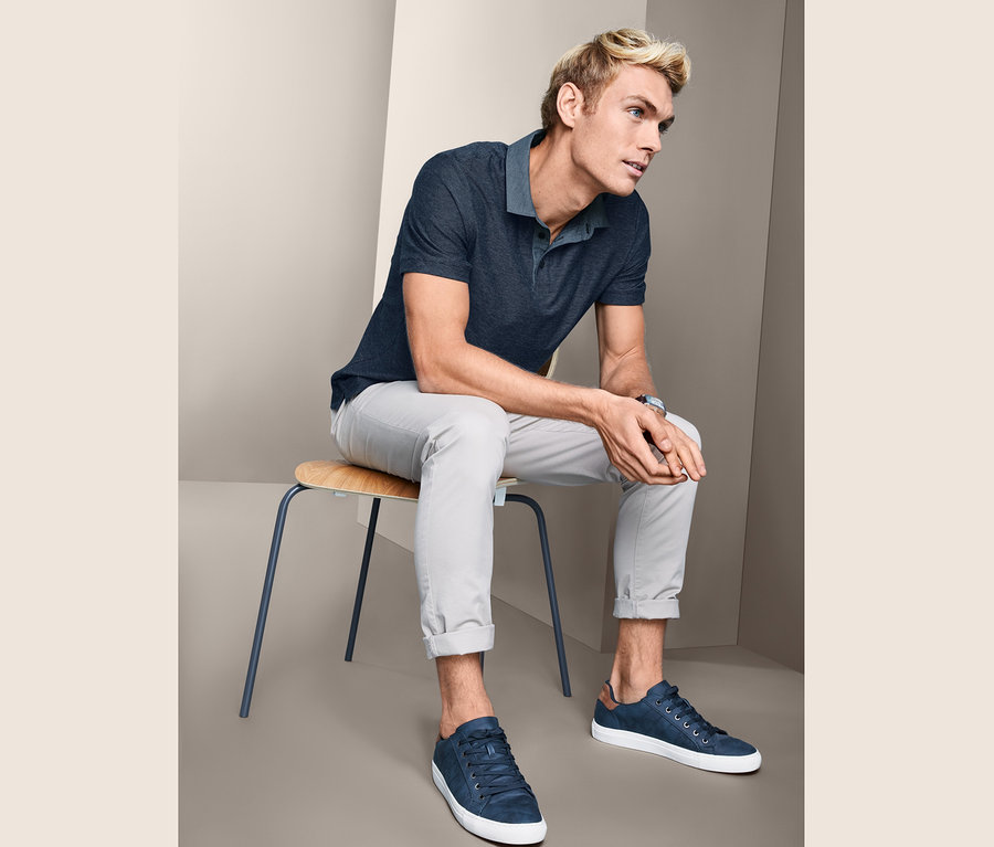 Mann sitzt auf einem Stuhl und trägt ein Jacquard-Poloshirt, Hose und Turnschuhe.