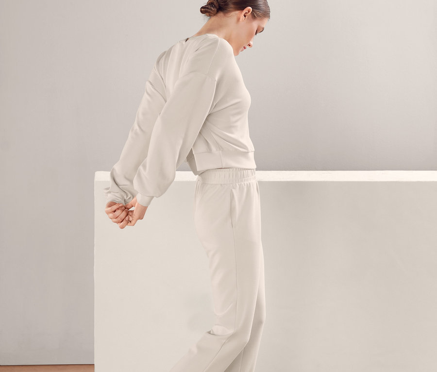 Eine Frau in einem beigen Trainingsanzug und Turnschuhen steht vor einer weißen Wand. Sie trägt einen extraweichen Sweatpullover.