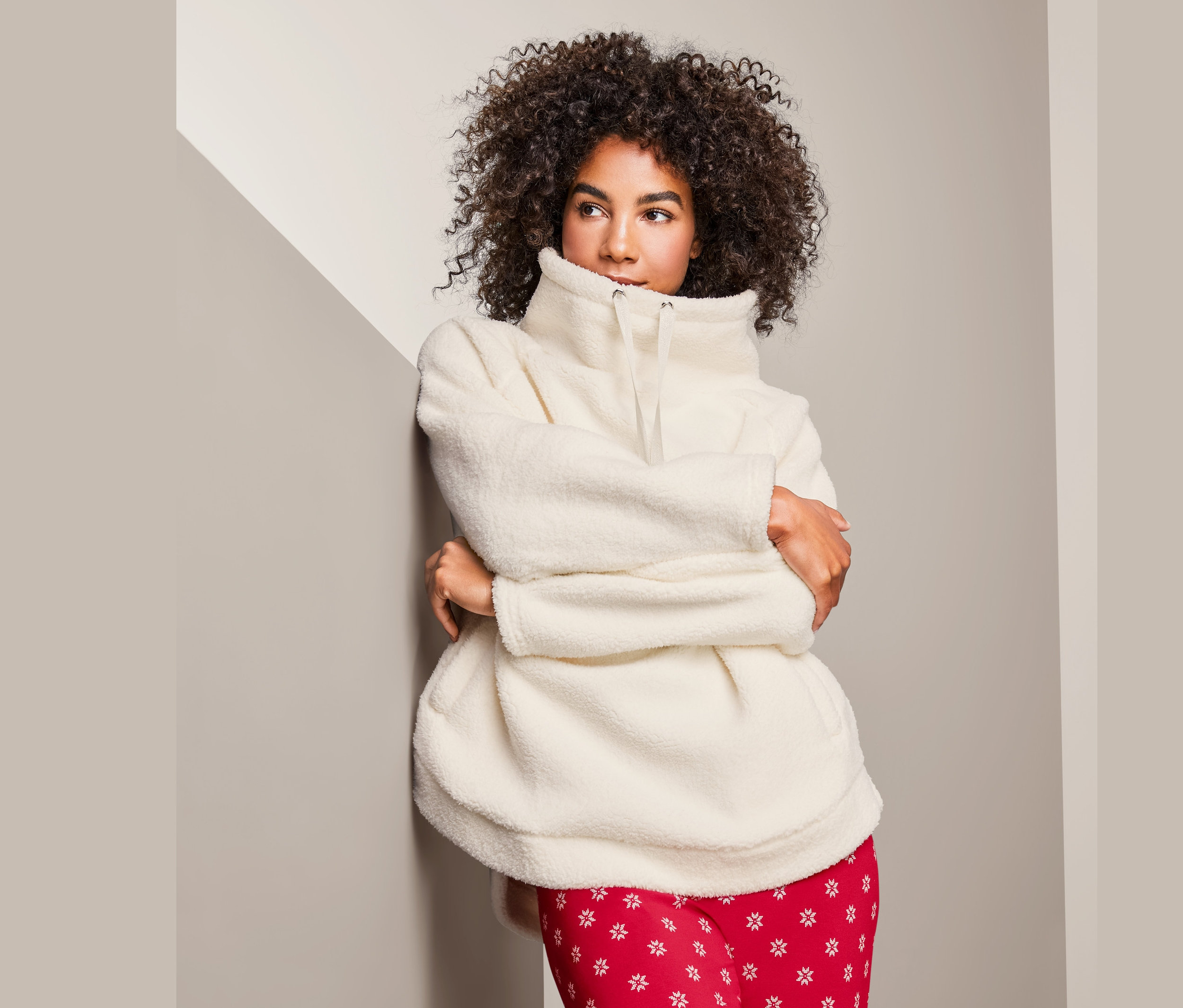 Frau mit lockigem Haar lehnt an der Wand und trägt einen Kuschel-Sweater und eine rote Hose.