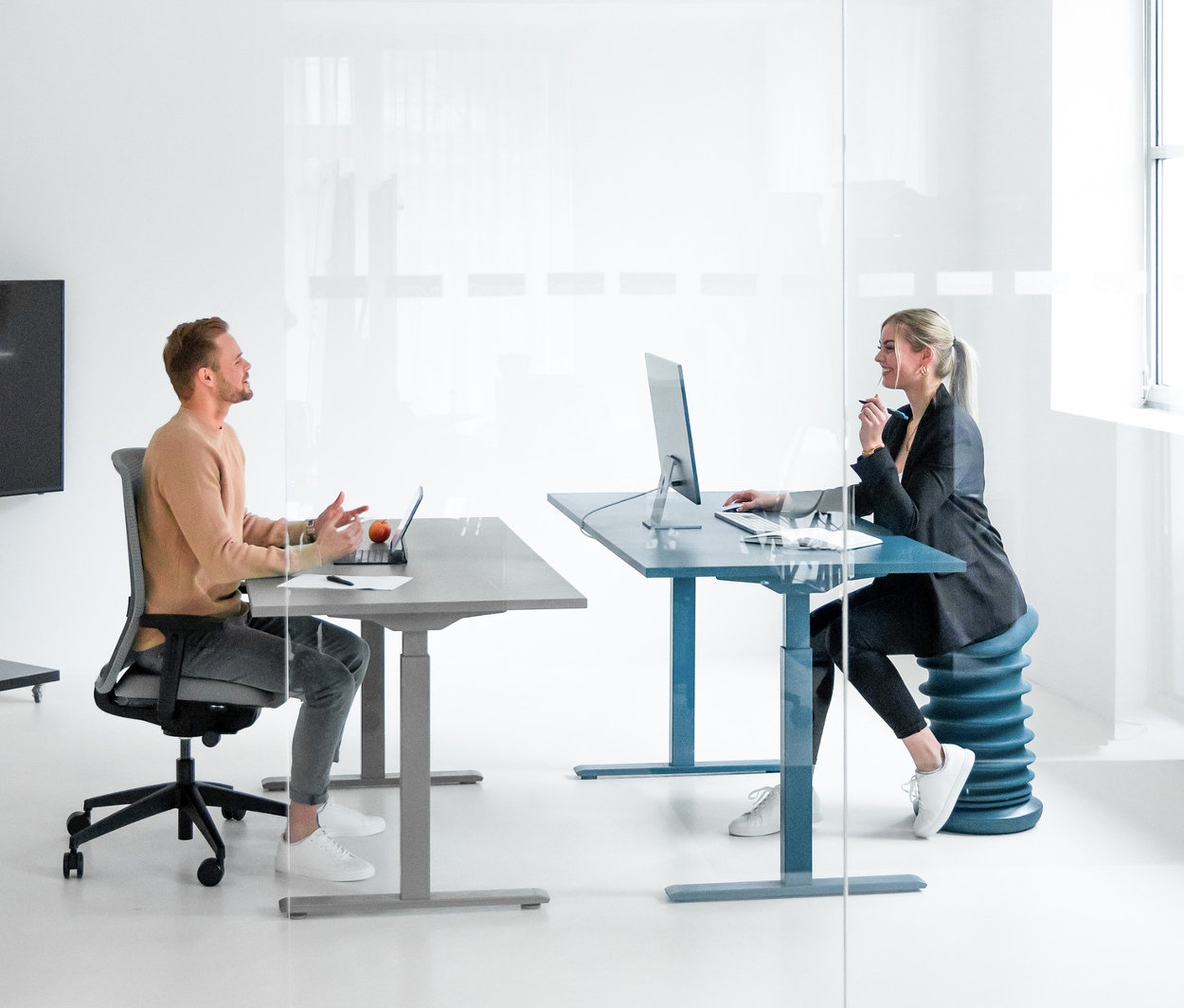 Mann sitzt an einem elektrisch verstellbaren Topstar Schreibtisch, arcticgrau, und Frau sitzt an einem elektrisch verstellbaren Topstar Schreibtisch, petrolblau.