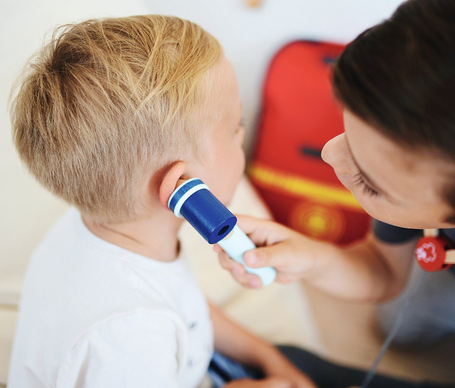 Eine Betreuerin misst die Temperatur eines kleinen Jungen mit einem Spielzeugthermometer. Im Hintergrund befindet sich ein kleiner small foot Notarzt-Rucksack.