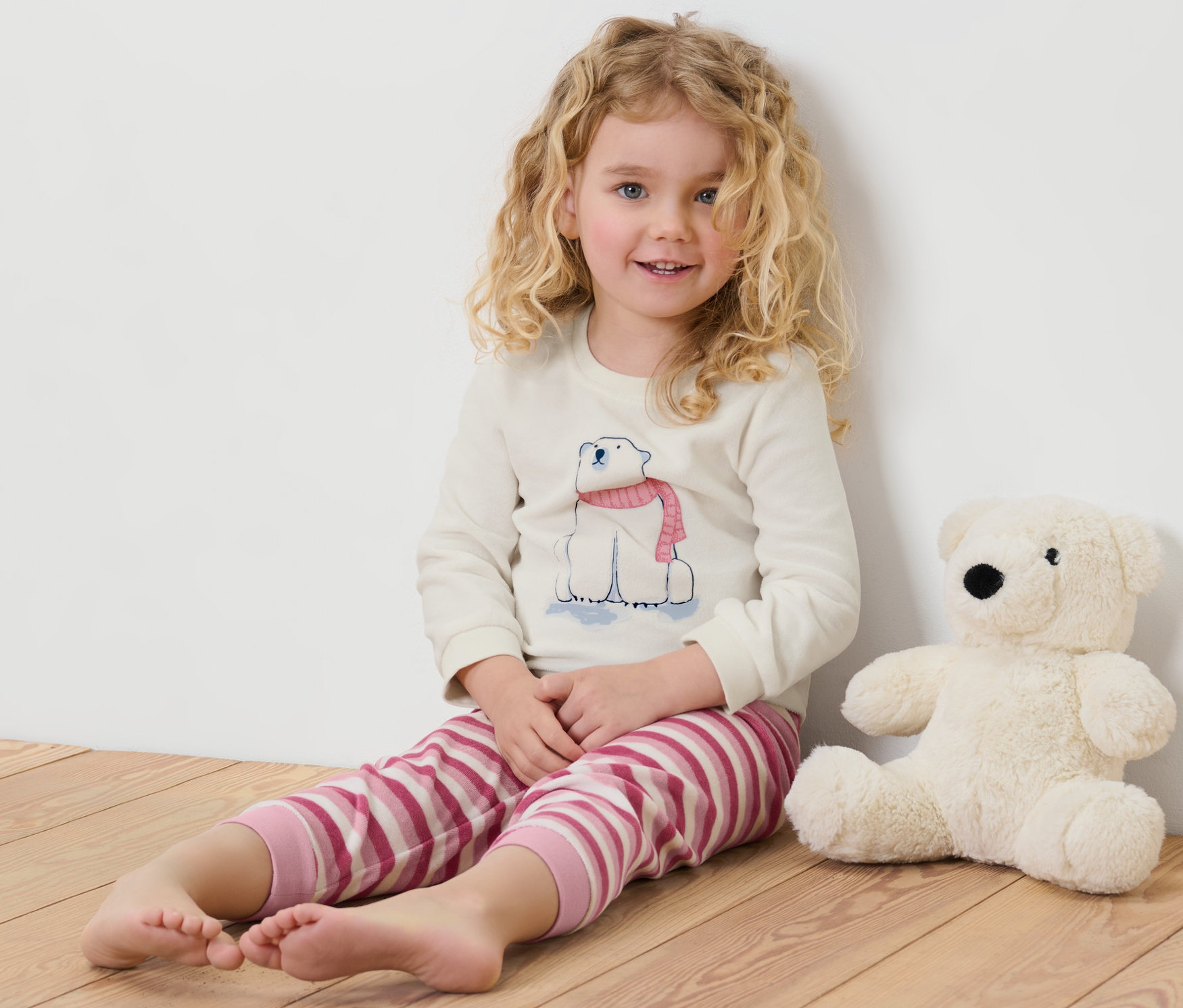 Mädchen sitzt auf dem Boden neben einem Stofftier-Eisbären. Sie trägt einen Kinder-Nicki-Pyjama mit einem Eisbärenmotiv.