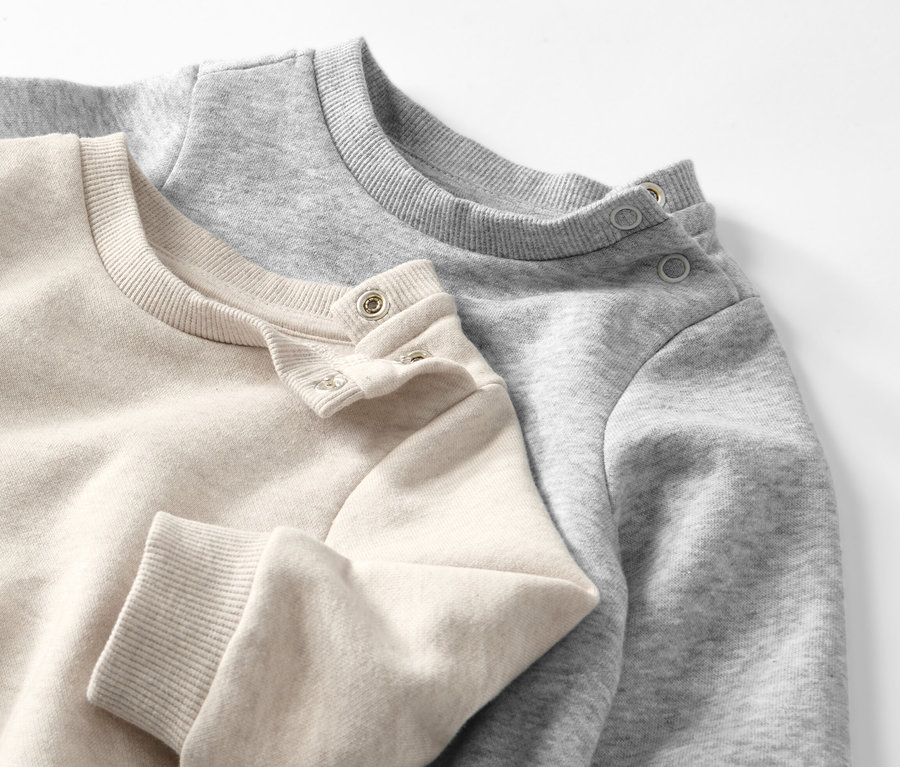 Zwei Baby-Sweatshirts mit angerauter Innenseite in Beige und Grau liegen übereinander.