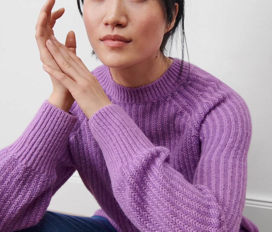 Eine Frau sitzt und trägt einen fliederfarbenen Strickpullover.