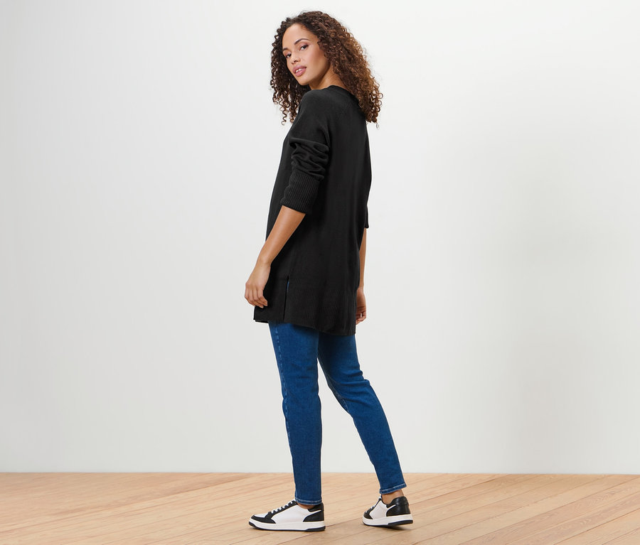 Frau blickt über die Schulter und trägt einen langen schwarzen Strickpullover, Jeans und Turnschuhe.