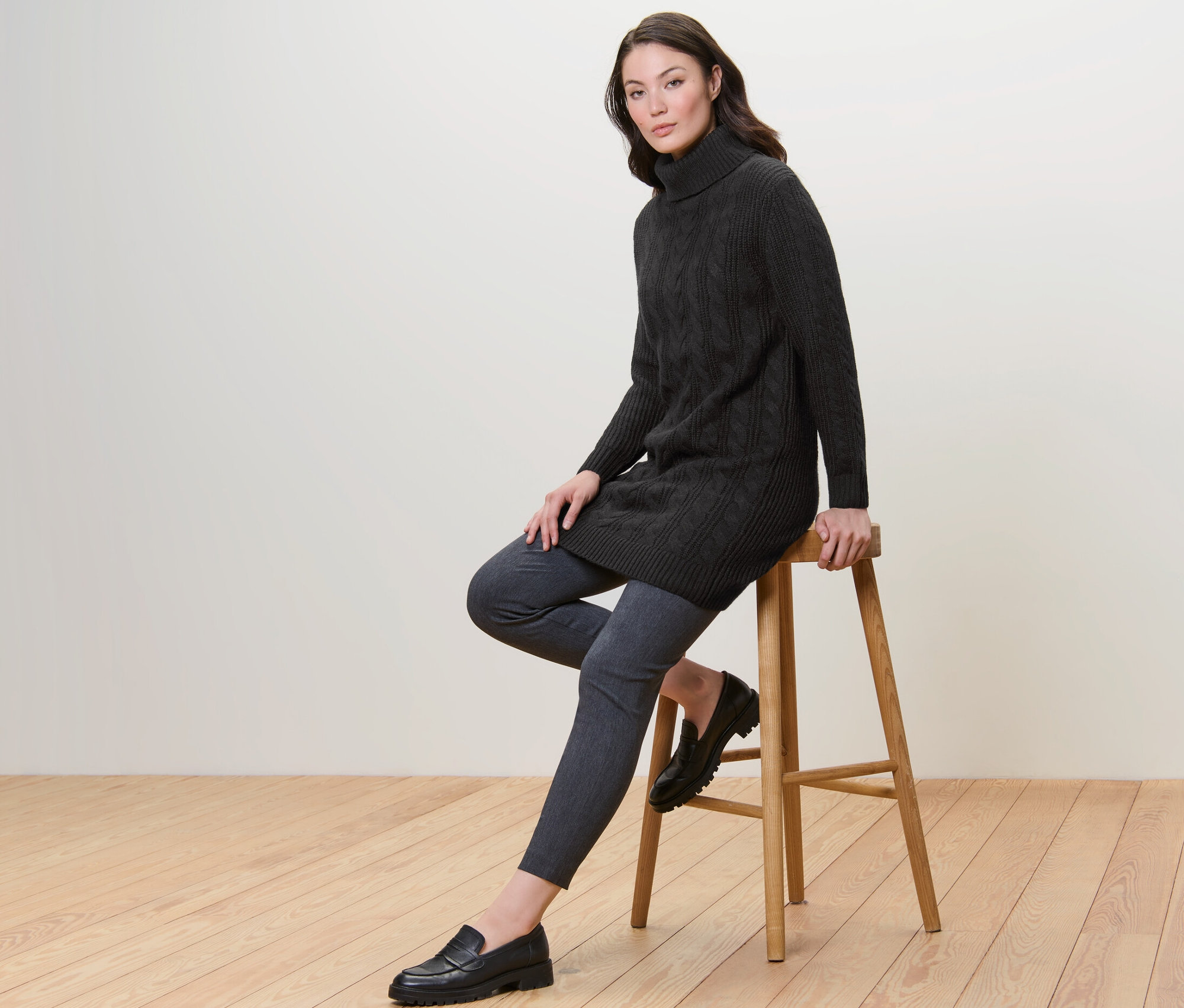 Frau sitzt auf einem Hocker und trägt einen langen, schwarzen Strickpullover mit Zopfmuster.