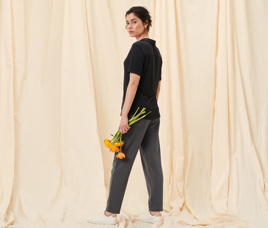 Frau blickt über die Schulter und hält orangefarbene Blumen. Sie trägt ein schwarzes Blusenshirt mit Knopfleiste und eine Joggpants mit Biese.