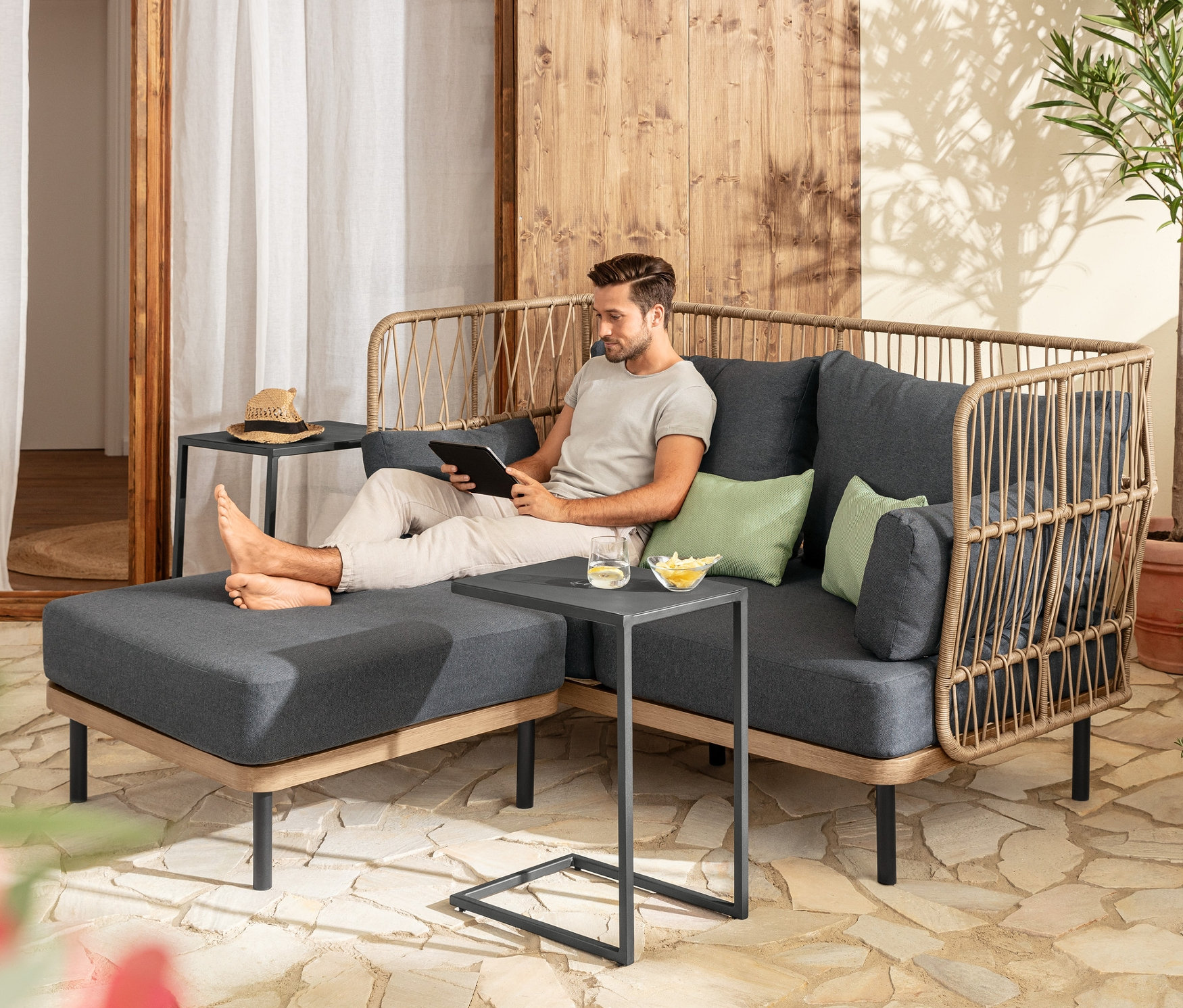 Mann sitzt auf einem Loungesofa mit Fußhocker und benutzt ein Tablet. Vorhanden sind 2 C-Tables und 2 Outdoor-Kissen.