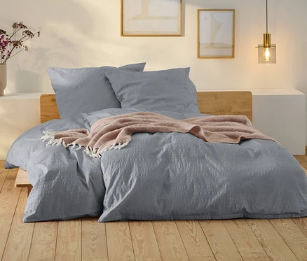 Graue Seersucker-Bettwäsche in Normalgröße und Übergröße auf einem Bett mit Holz Kopfteil. Auf dem Bett liegt eine rosafarbene Decke.