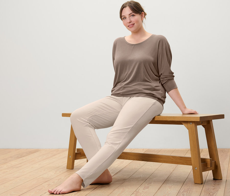Frau sitzt auf einer Bank, trägt ein taupefarbenes Sport-und-Yogashirt und eine beige Sweathose.