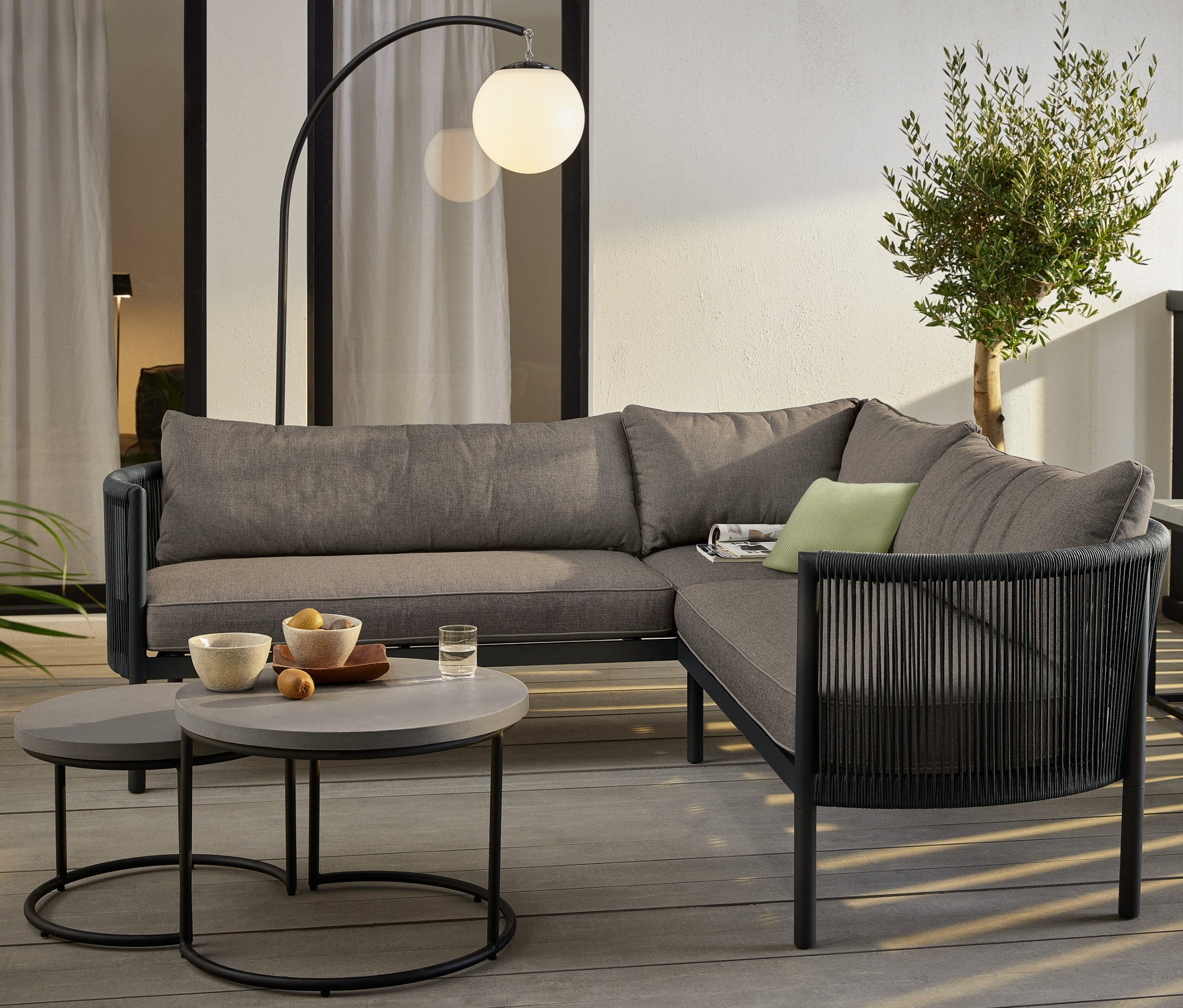 Eine Lounge-Ecke »Jara« mit Textilgeflecht steht auf einer Terrasse, neben 2 Couchtischen mit Beton-Tischplatte. Eine LED-Akku-Bogenleuchte steht neben der Lounge.