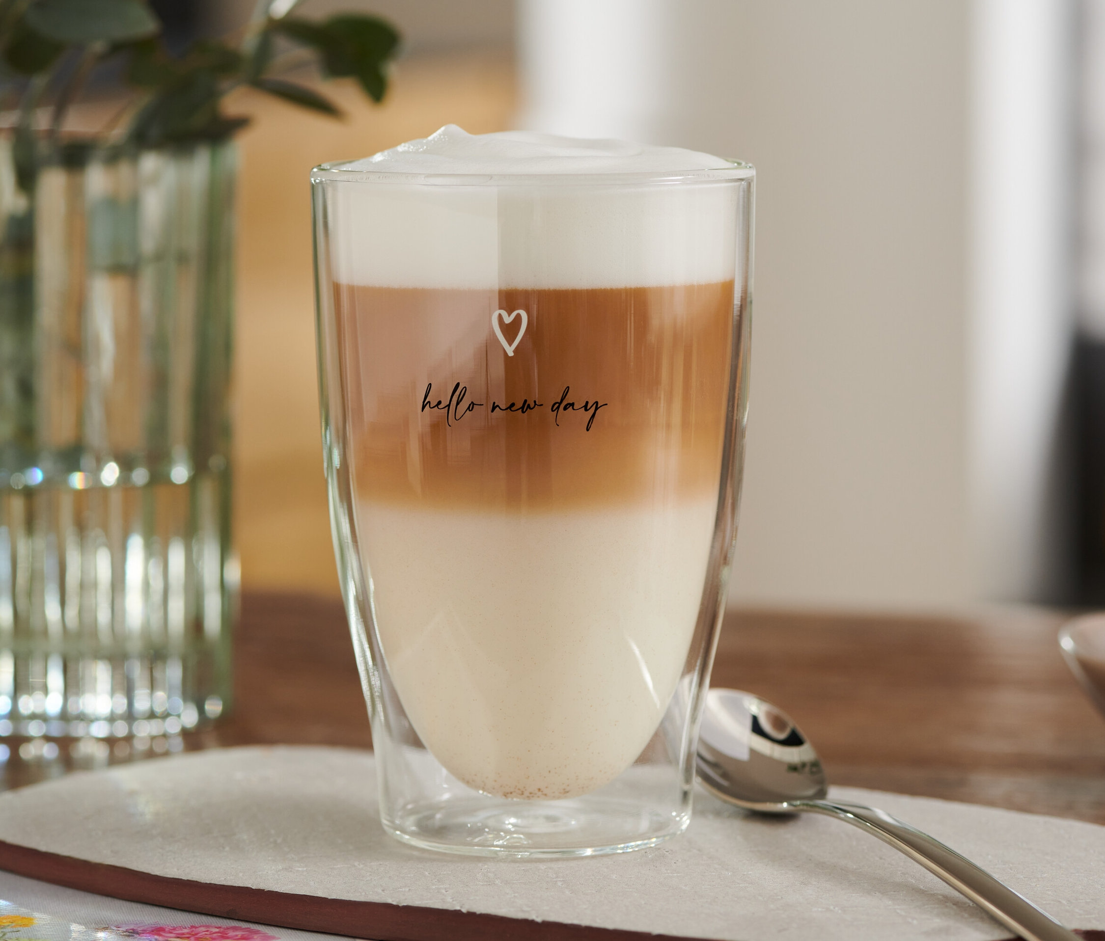 Ein Latte Macchiato Glas, bedruckt mit Herzchen, gefüllt mit Milch-, Kaffee- und Schaumschichten.