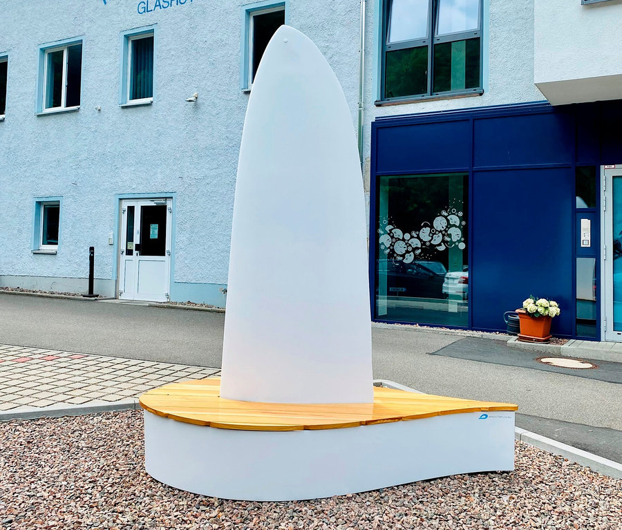 Außenaufnahme einer weißen Wings for Living Bank »Sydney« mit Holzsitzen auf einer Kiesfläche, im Hintergrund ein Gebäude.