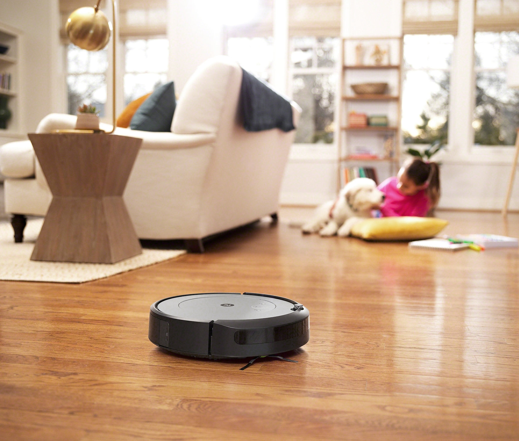 Ein schwarzer iRobot Saugroboter »Roomba i1« fährt auf einem Holzboden. Im Hintergrund liegen ein Mädchen und ein Hund.