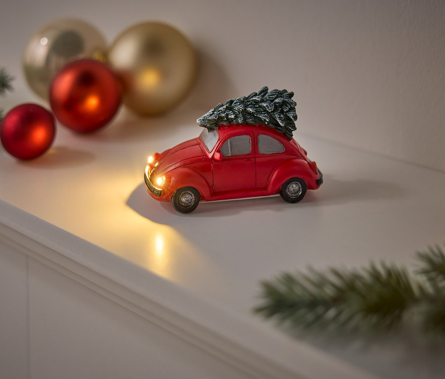 Ein rotes Spielzeugauto mit einem Weihnachtsbaum auf dem Dach steht auf einem weißen Regal, zusammen mit roten und goldenen Weihnachtskugeln und einem Tannenzweig.