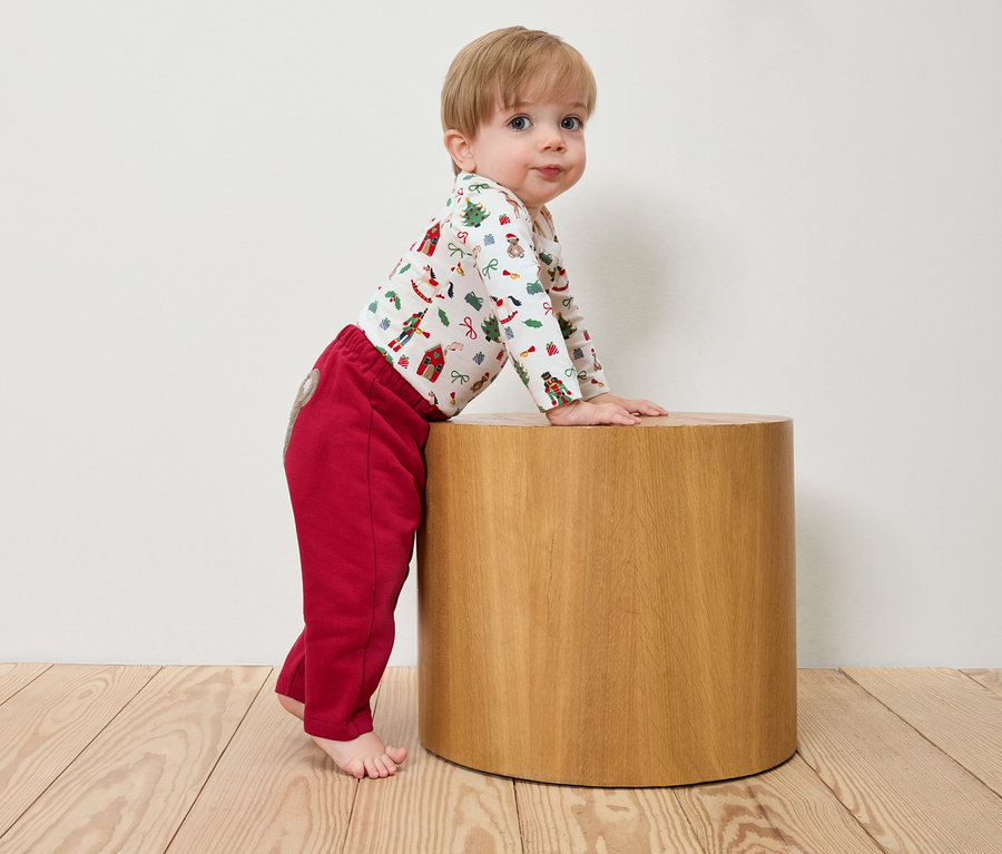 Kleinkind lehnt an einem Holzhocker, trägt einen weihnachtlichen Body und rote Hosen.