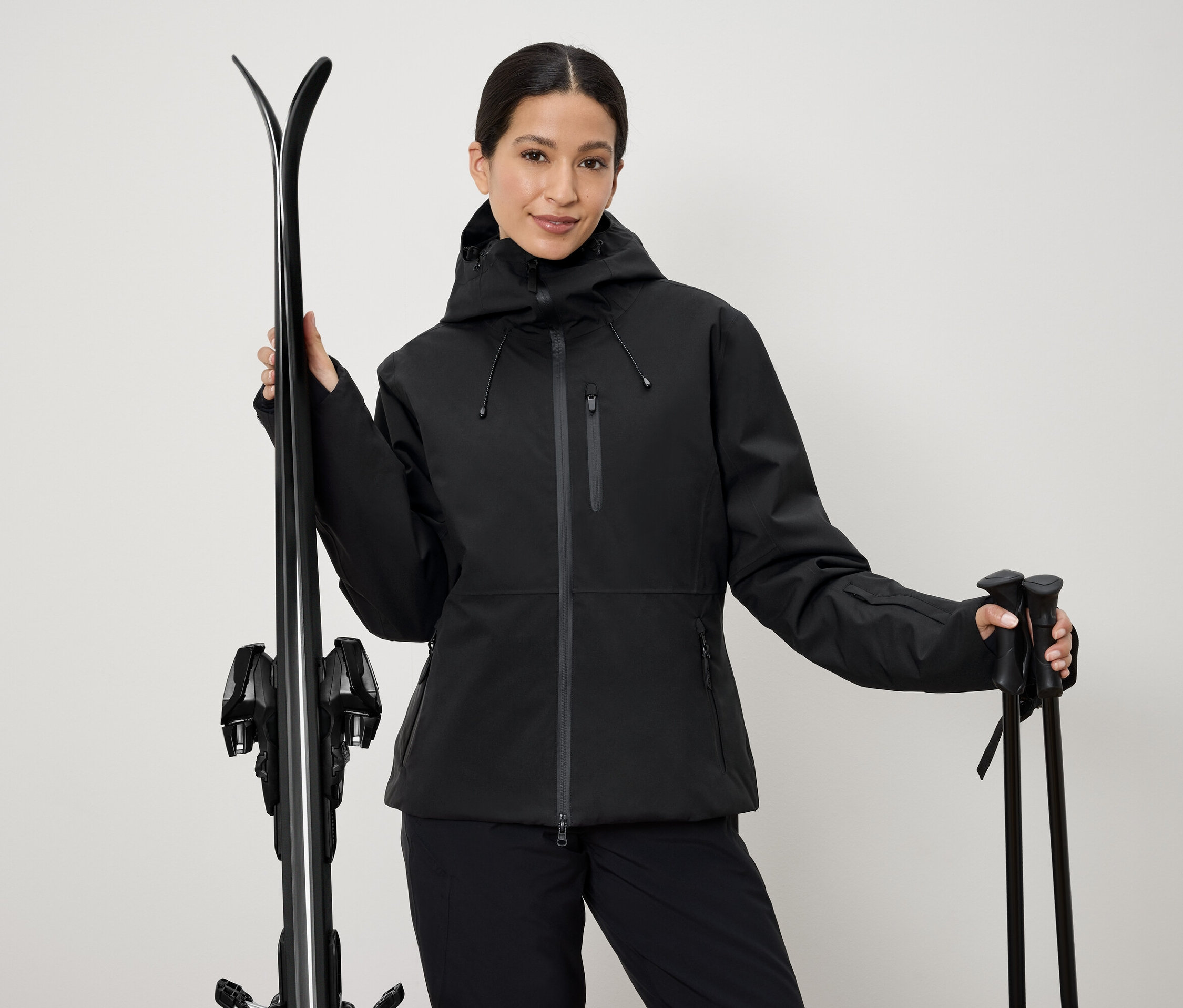 Frau in schwarzer Skijacke hält Ski und Stöcke.