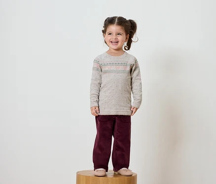 Lächelndes kleines Mädchen mit braunen Haaren in Zöpfen steht auf einem Holzsockel. Sie trägt einen beige Pullover und bordeauxrote Hosen.
