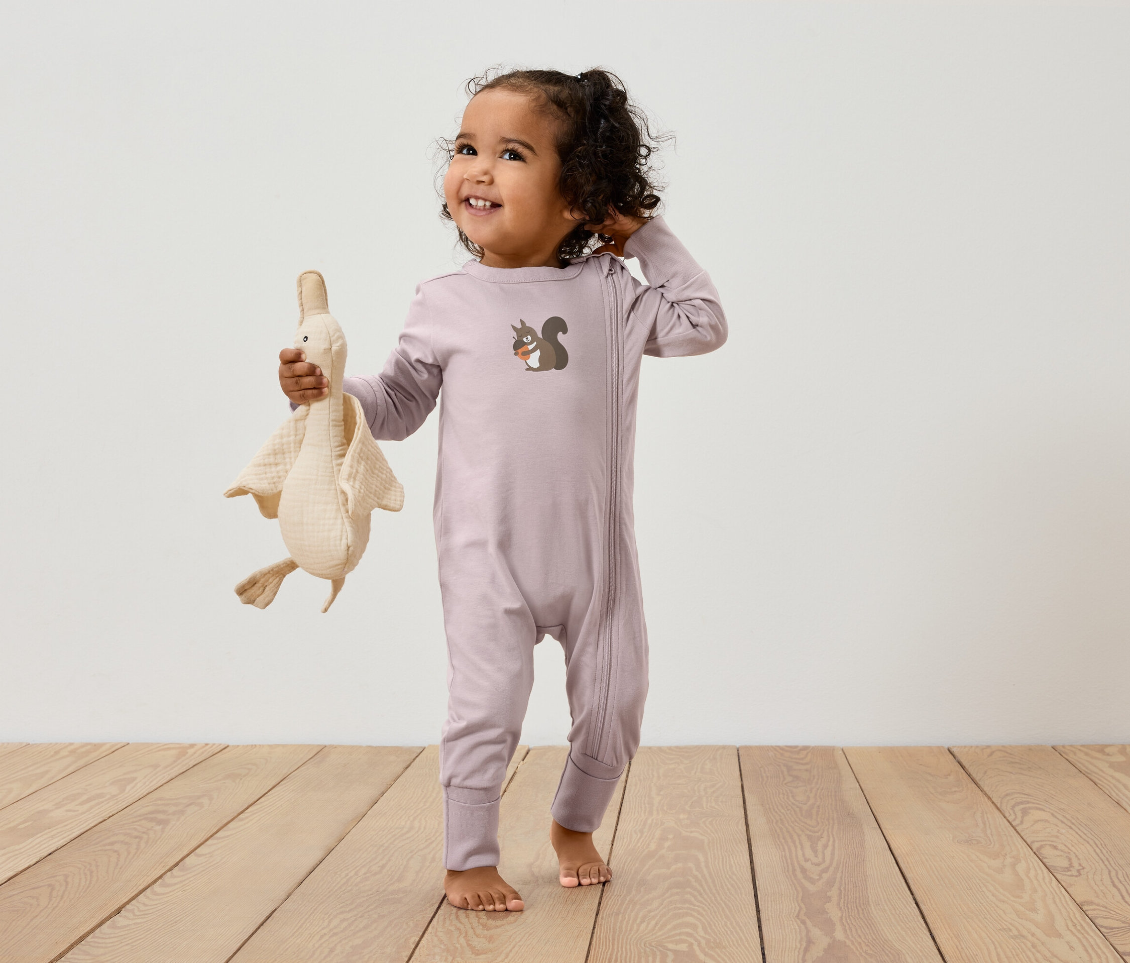 Ein Kind in einem rosa Overall mit Reißverschluss und einem Eichhörnchenbild hält ein Plüschtier einer Gans.