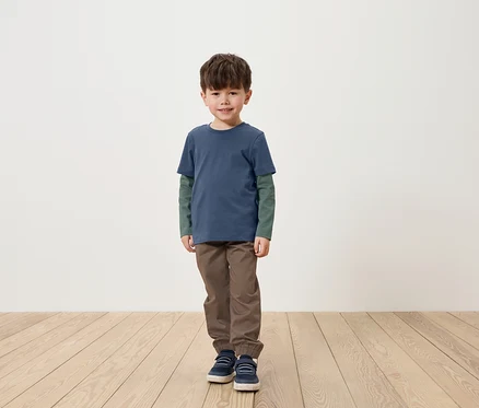 Ein Junge steht auf einem Holzboden und trägt ein blaues T-Shirt mit langen Ärmeln, eine braune Hose und blaue Schuhe.
