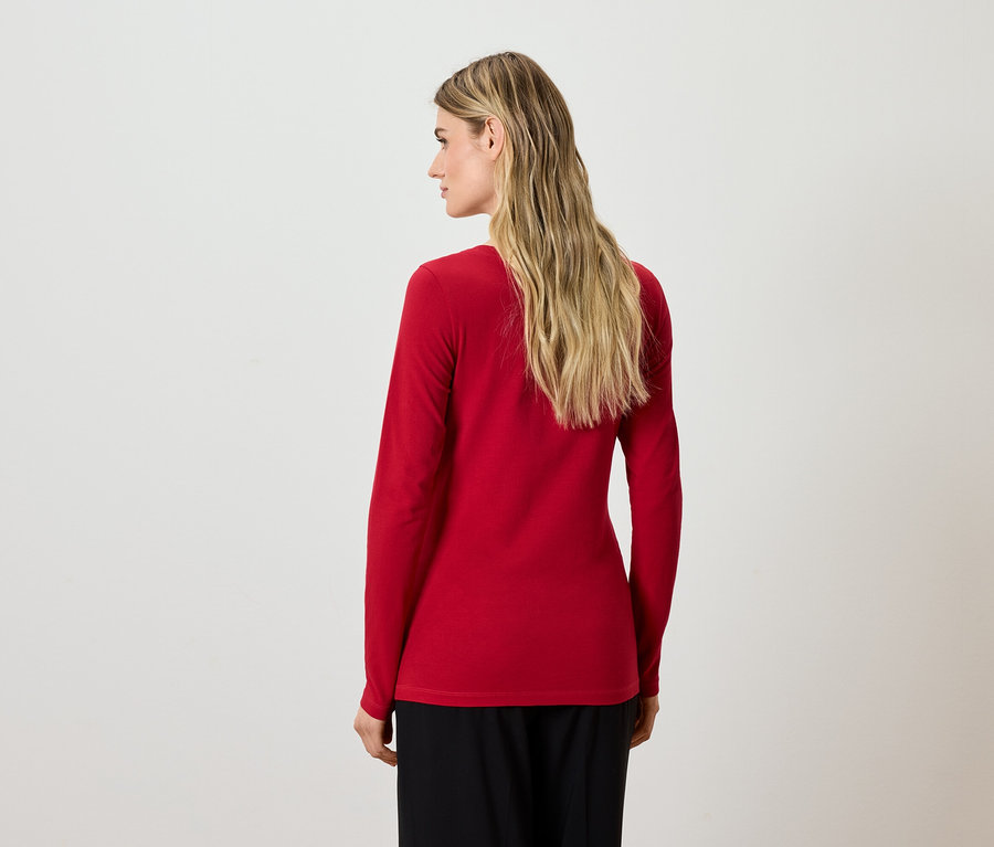 Blonde Frau steht mit dem Rücken zur Kamera, trägt eine rote Bluse mit langen Ärmeln und eine schwarze Hose.