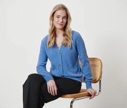 Blonde Frau sitzt auf einem Stuhl und trägt einen blauen Cardigan und eine schwarze Hose.