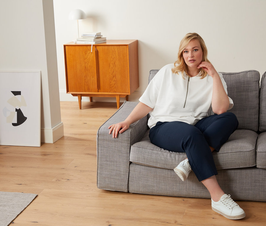 Frau sitzt auf einem grauen Sofa und trägt ein weißes Oversized-Sweatshirt, eine blaue 7/8-Stretchhose und Canvas-Sneaker.