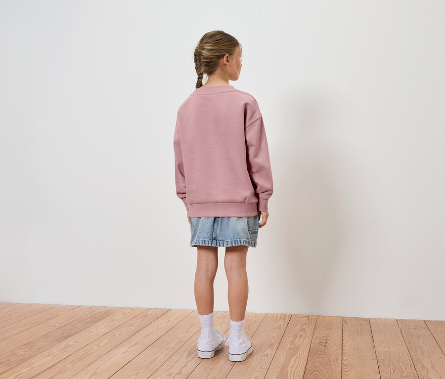 Mädchen mit Zopf trägt einen rosafarbenen Pullover und einen Jeansrock, von hinten gesehen.