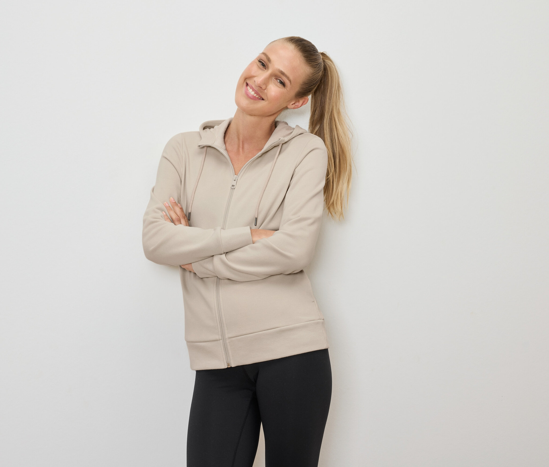 Lächelnde blonde Frau mit Pferdeschwanz posiert mit verschränkten Armen in einer beige Kapuzenjacke mit Reißverschluss und schwarzen Leggings.