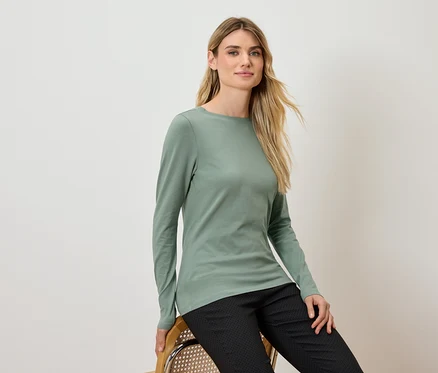 Blonde Frau sitzt auf einem Stuhl, trägt ein grünes Langarmshirt und eine schwarze Hose.