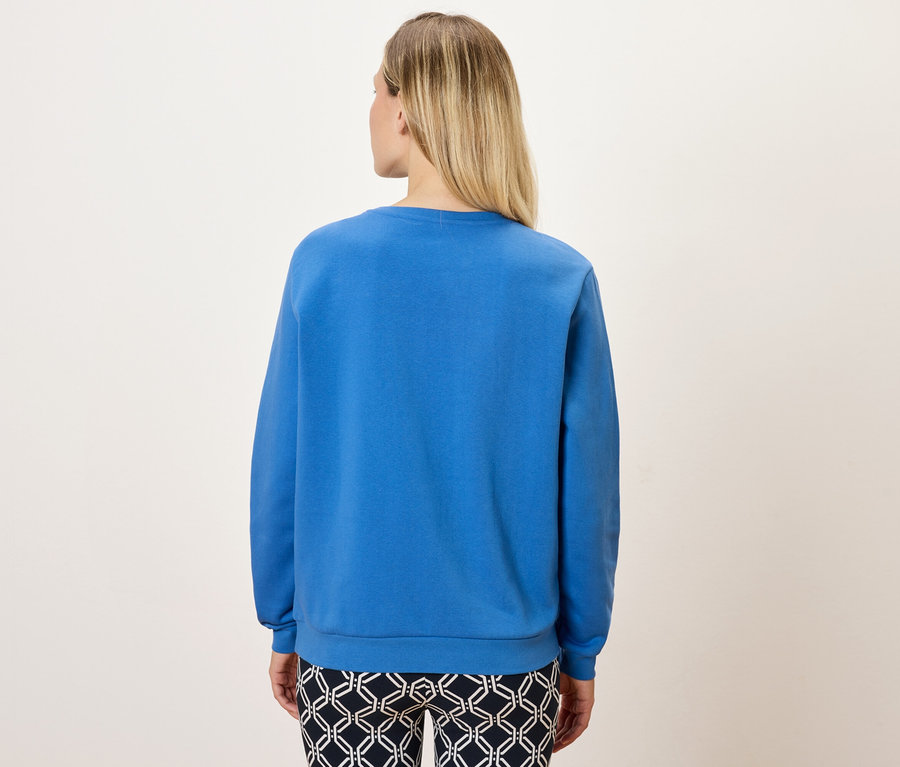 Rückansicht einer blonden Frau mit blauem Sweatshirt und gemusterter Hose.