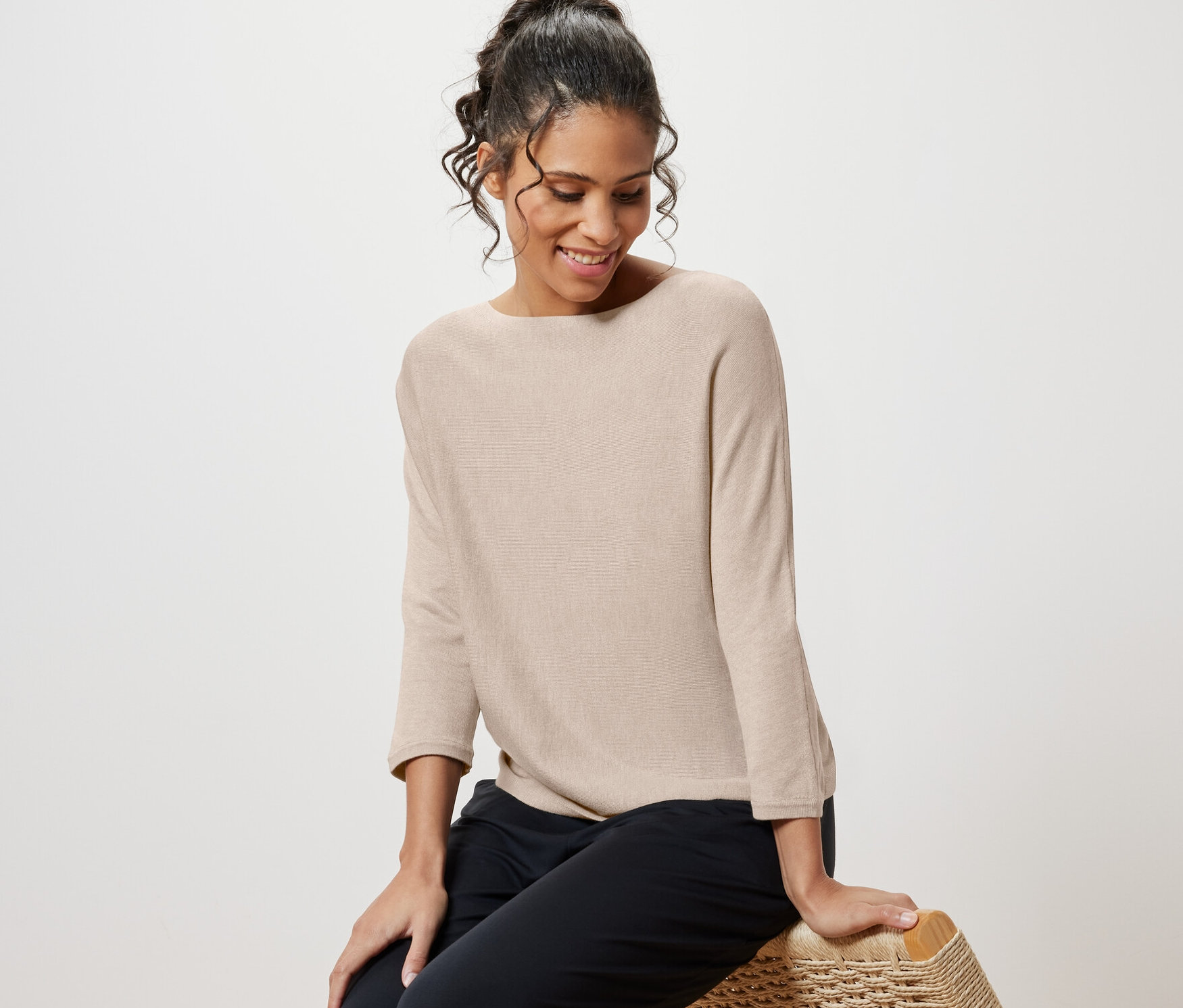 Frau sitzt auf Korbstuhl und trägt einen beige melierten Feinstrickpullover.