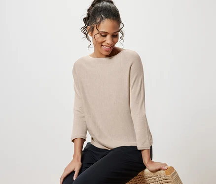 Frau sitzt auf Korbstuhl und trägt einen beige melierten Feinstrickpullover.