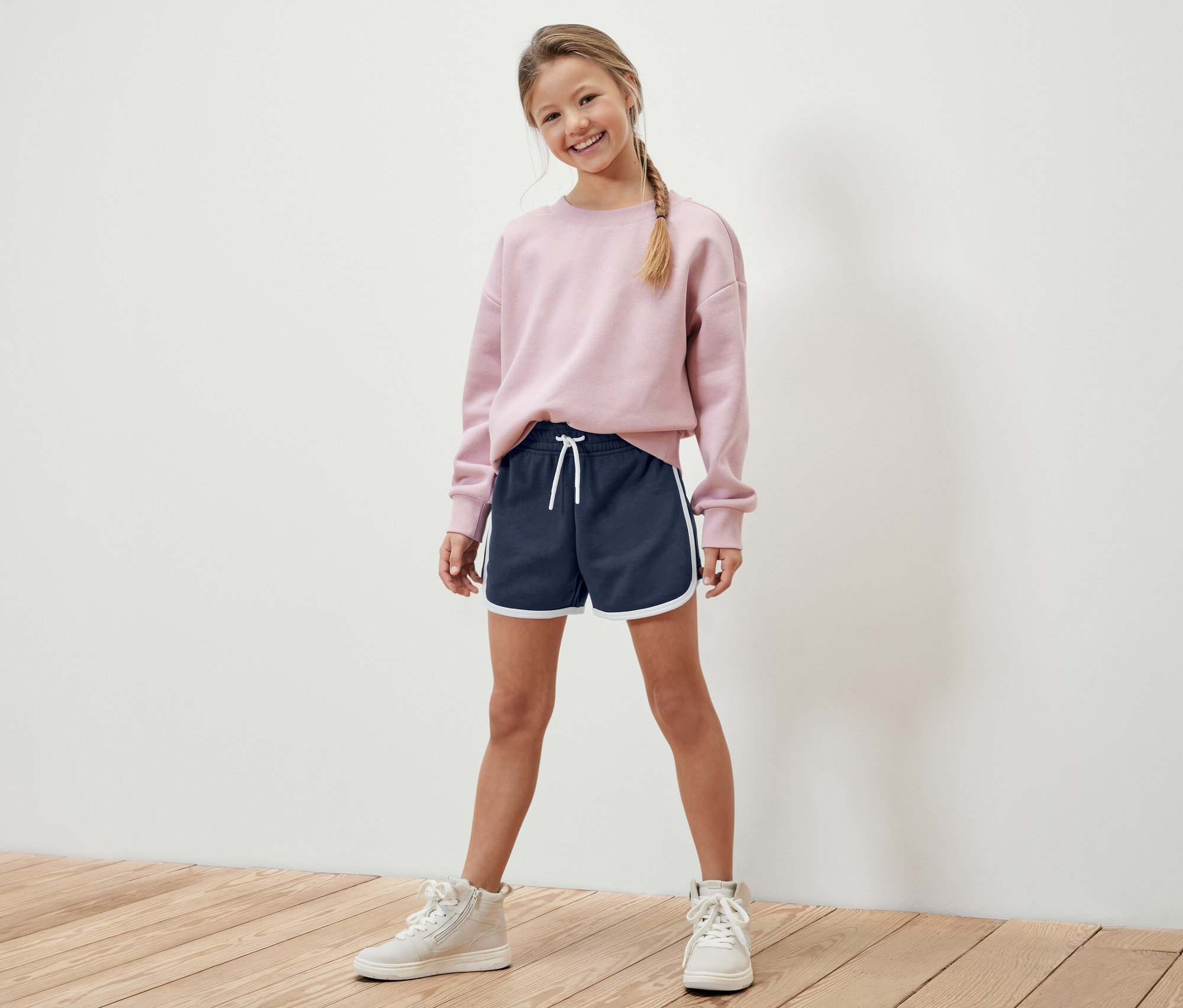 Mädchen posiert in rosa Sweatshirt und dunkelblauen Sweatshorts mit weißem Saum. Sie trägt 2 Kinder-Sweatshorts.