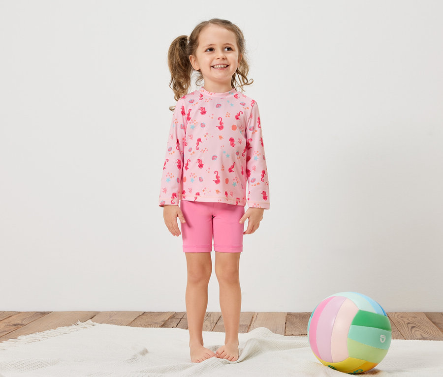 Ein Mädchen mit Zöpfen trägt ein rosa Langarmshirt mit Seepferdchenmuster und pinke Shorts. Ein bunter Ball liegt auf einem weißen Teppich.