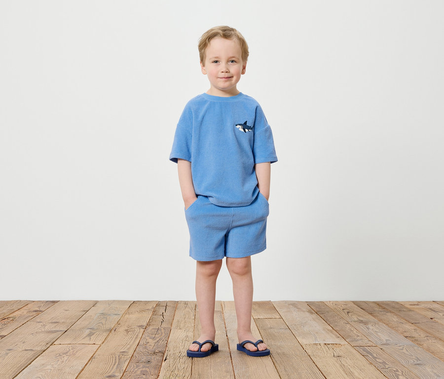 Ein Junge steht vor einer weißen Wand, gekleidet in ein blaues T-Shirt mit Hai-Motiv und blaue Shorts, dazu trägt er blaue Flip-Flops.