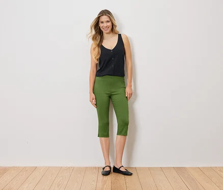 Frau steht vor einer weißen Wand und trägt ein schwarzes Top, eine grüne Caprihose und schwarze Ballerinas.