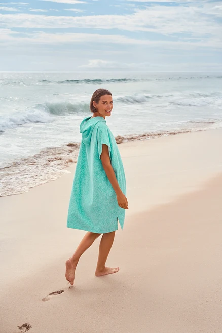 Frau in türkisfarbenem Poncho mit Kapuze, die barfuß am Strand entlanggeht und über die Schulter blickt.