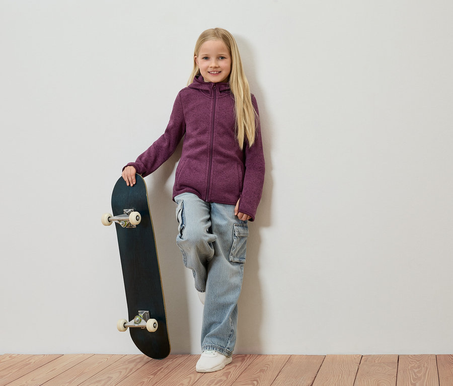 Mädchen mit blonden Haaren lehnt an einer Wand, hält ein Skateboard und trägt eine lila Fleecejacke, Jeans und weiße Schuhe.
