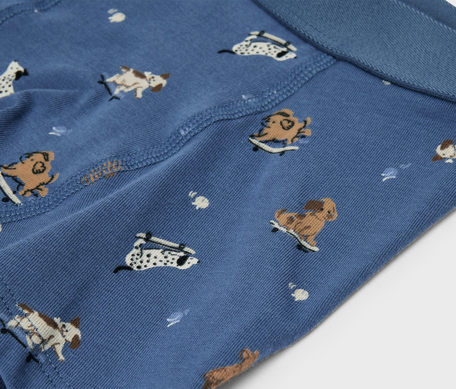 Nahaufnahme von blauem Stoff mit einem Muster aus verschiedenen Cartoon-Hunden auf Skateboards. Der Stoff zeigt Nähte und einen blauen Bund.
