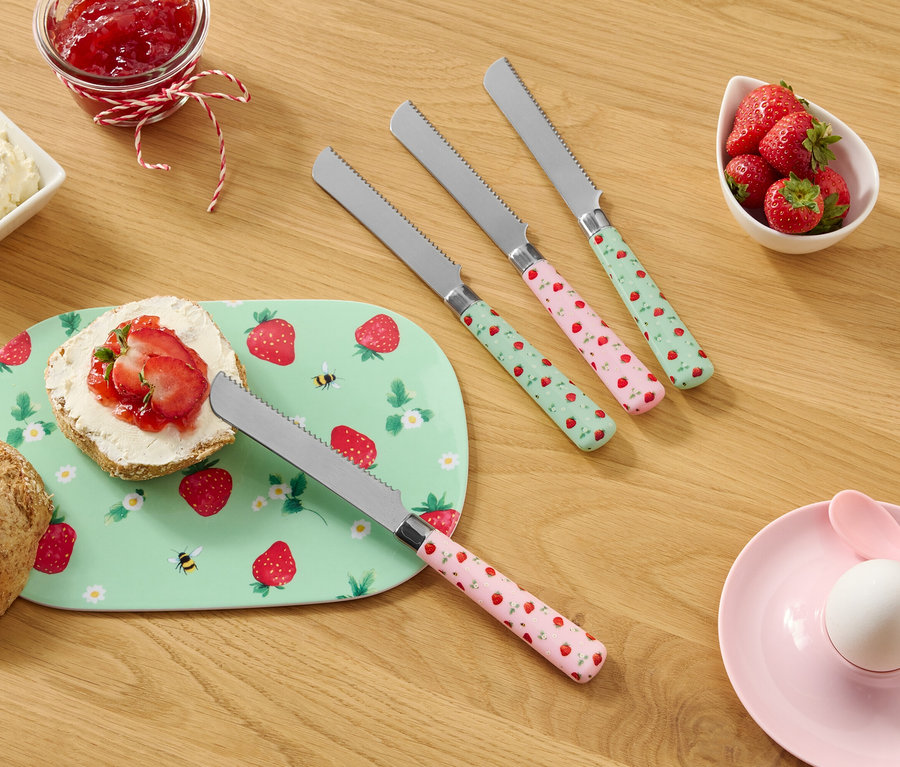 Auf einem Holztisch befinden sich ein Brötchen, ein Teller mit Erdbeermuster, Frischkäse, Marmelade, Erdbeeren und vier Messer mit Erdbeermuster.