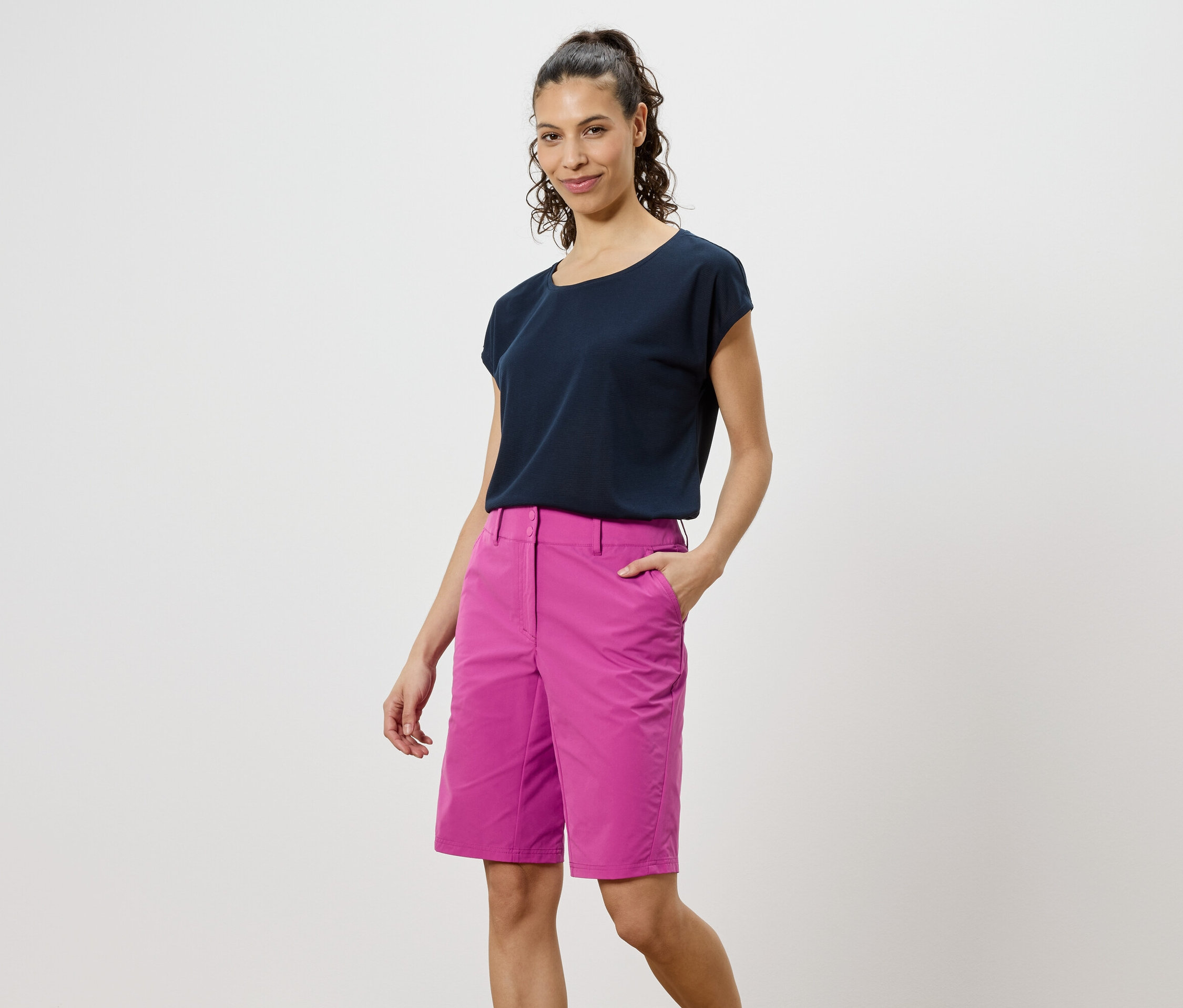 Frau mit dunklem T-Shirt und pinken Shorts steht vor einem weißen Hintergrund.